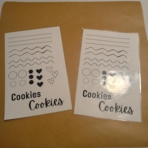 May include: Two white cards with black line drawings and the word "Cookies" in cursive. The cards have various line patterns, circles, dots, and hearts. One card is in a clear plastic sleeve.