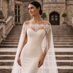 Könnte beinhalten: Ein cremefarbenes Hochzeitskleid mit herzförmigem Ausschnitt und einem transparenten, mit Spitze besetzten Umhang. Das Kleid ist figurbetont und bodenlang. Der Hintergrund zeigt ein Steingebäude und eine Treppe.