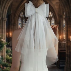 Puede incluir: Un velo de novia de tul blanco con un lazo grande y detalles de perlas. El velo es de varias capas y cae por la espalda. El lazo está sujeto al cabello. Accesorios de boda y novia.