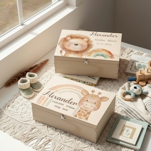 Könnte beinhalten: Zwei hölzerne Andenkenboxen mit Illustrationen eines Löwen und einer Giraffe, jeweils mit einem Regenbogen-Design. Die Boxen sind hellbraun mit silbernen Verschlüssen. Eine Box trägt den Namen "Alexander" und Geburtsdetails.