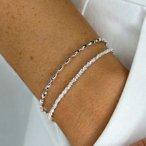 May include: Two silver bracelets are shown on an arm. The upper bracelet has a series of oval links. The lower bracelet has a textured, sparkling appearance. The bracelets are set against a white background.