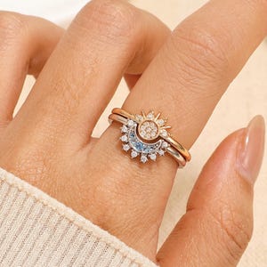 May include: A set of two rings, one gold and one silver, featuring celestial designs. The gold ring has a sunburst design with small, clear stones. The silver ring has a crescent moon design with clear and blue stones.