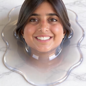 Può includere: Il volto sorridente di una persona è circondato da una sostanza trasparente, simile a un liquido, creando un effetto visivo unico. La persona ha capelli e occhi castani e indossa piccoli orecchini d'oro. Lo sfondo è una superficie di marmo bianco.