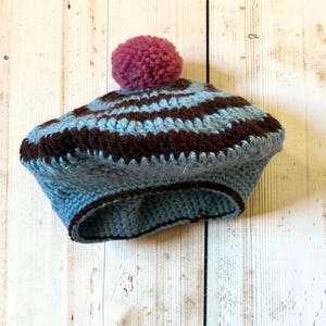 May include: A crocheted beret featuring alternating horizontal stripes of light blue and dark brown yarn. The hat is topped with a single, round, textured pom-pom in a dusty rose pink colour, and the base is finished with a dark brown trim.