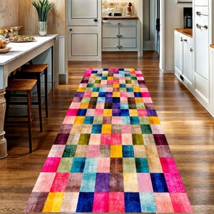 May include: A long rectangular runner rug featuring a vibrant grid pattern of multicolored squares in shades of pink, blue, yellow, green, and purple. The rug is positioned on a wooden floor in a kitchen setting with white cabinets and stools.