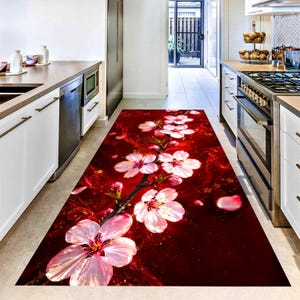 May include: A rectangular kitchen rug featuring a vibrant floral design. The rug showcases a deep red background with a branch of pink and white blossoms. Petals are scattered around the flowers, creating a visually appealing pattern. The rug is designed to fit a kitchen hallway.