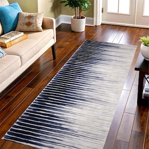 May include: A rectangular rug featuring an abstract design in navy blue and white. The pattern comprises horizontal lines that transition from dark blue to white. The rug is positioned on a wooden floor, adjacent to a beige sofa.