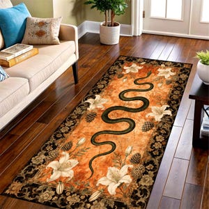 May include: A rectangular rug with an orange background and a black border. The rug features a large, winding green snake design, white lilies, and pinecones. The rug is placed on a hardwood floor.