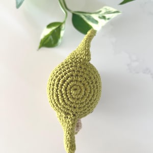 Puede incluir: Un art&iacute;culo de ganchillo hecho a mano, de color verde claro, con un dise&ntilde;o en espiral en la parte superior. El art&iacute;culo tiene una parte superior puntiaguda y una peque&ntilde;a base beige. El fondo incluye hojas abigarradas verdes y blancas.