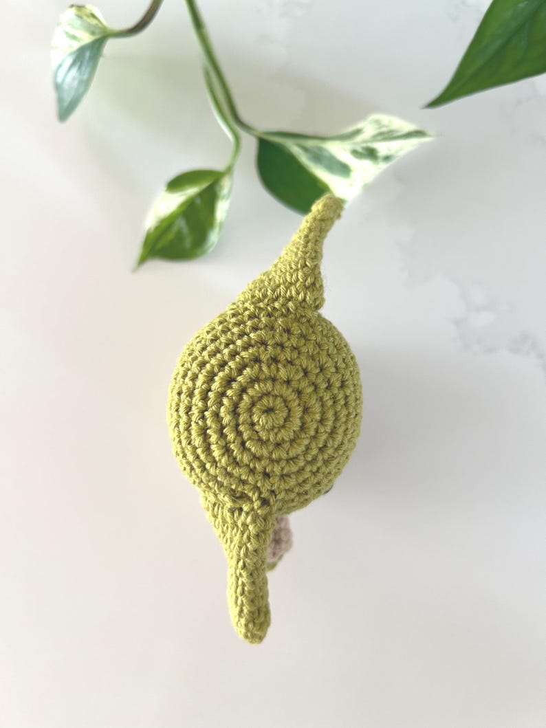 Puede incluir: Un art&iacute;culo de ganchillo hecho a mano, de color verde claro, con un dise&ntilde;o en espiral en la parte superior. El art&iacute;culo tiene una parte superior puntiaguda y una peque&ntilde;a base beige. El fondo incluye hojas abigarradas verdes y blancas.