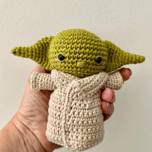Puede incluir: Mu&ntilde;eca de ganchillo hecha a mano de un personaje con orejas grandes y ojos negros. La mu&ntilde;eca es verde con una t&uacute;nica beige. La mu&ntilde;eca se sostiene en una mano.
