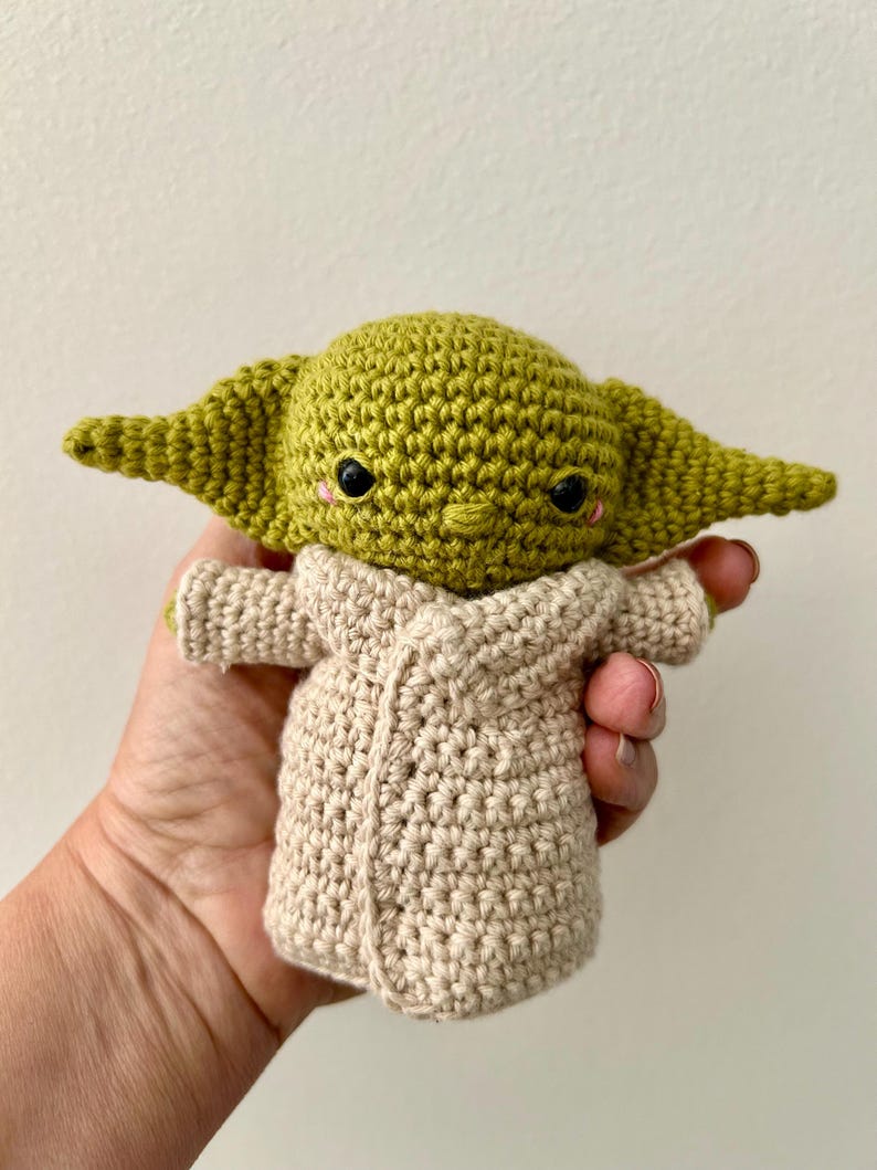 Puede incluir: Mu&ntilde;eca de ganchillo hecha a mano de un personaje con orejas grandes y ojos negros. La mu&ntilde;eca es verde con una t&uacute;nica beige. La mu&ntilde;eca se sostiene en una mano.