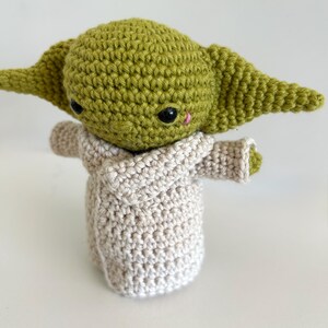 Puede incluir: Mu&ntilde;eco de ganchillo hecho a mano de un personaje con orejas grandes y cabeza verde claro. El mu&ntilde;eco lleva una t&uacute;nica beige y tiene ojos negros y una peque&ntilde;a nariz rosa. El tema del mu&ntilde;eco es Star Wars.