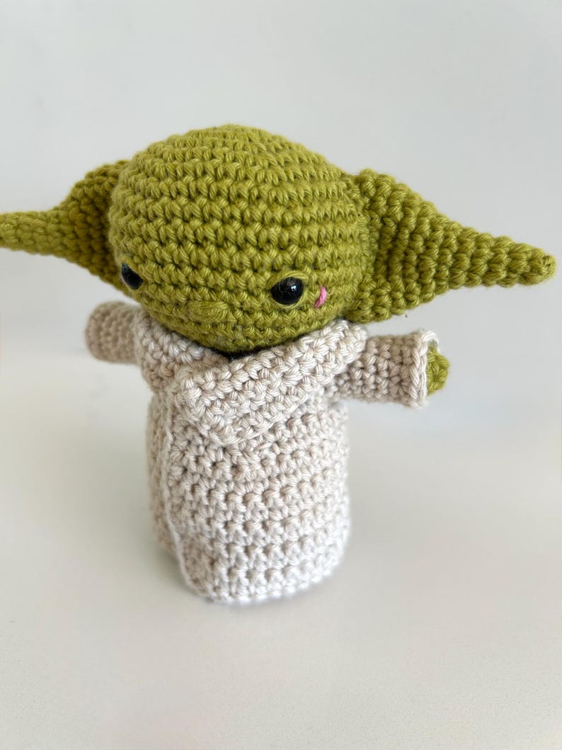 Puede incluir: Mu&ntilde;eco de ganchillo hecho a mano de un personaje con orejas grandes y cabeza verde claro. El mu&ntilde;eco lleva una t&uacute;nica beige y tiene ojos negros y una peque&ntilde;a nariz rosa. El tema del mu&ntilde;eco es Star Wars.