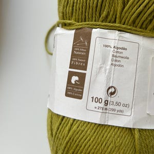 Puede incluir: Primer plano de un ovillo de hilo de algod&oacute;n verde oliva. La etiqueta indica que es 100% algod&oacute;n, 100 g y aproximadamente 275 metros. La etiqueta tambi&eacute;n incluye texto en portugu&eacute;s, alem&aacute;n e ingl&eacute;s.