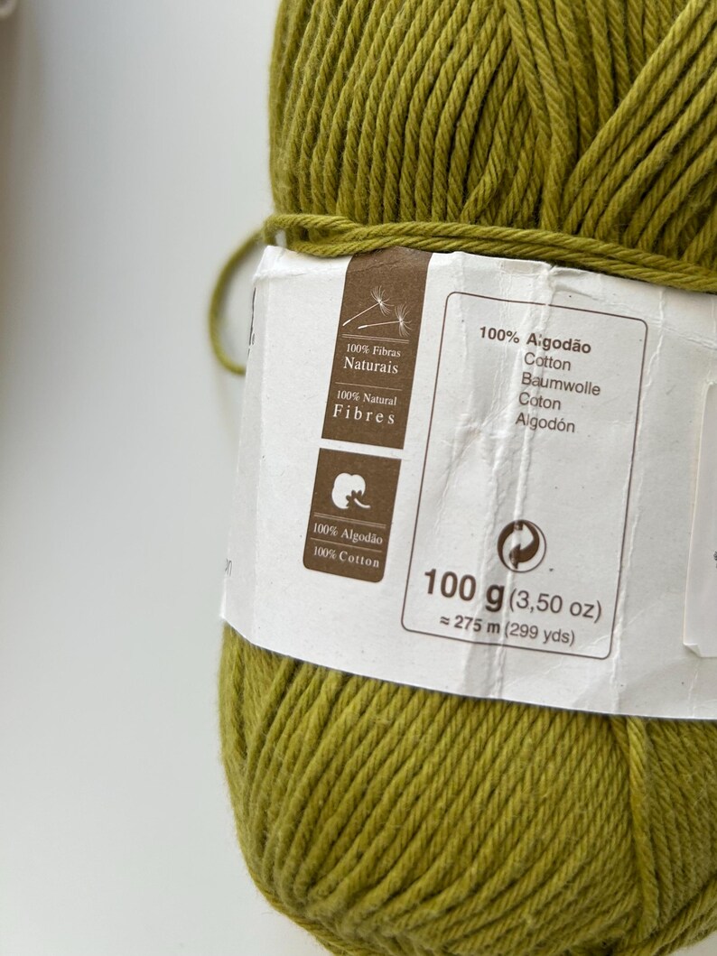 Puede incluir: Primer plano de un ovillo de hilo de algod&oacute;n verde oliva. La etiqueta indica que es 100% algod&oacute;n, 100 g y aproximadamente 275 metros. La etiqueta tambi&eacute;n incluye texto en portugu&eacute;s, alem&aacute;n e ingl&eacute;s.