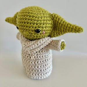 Puede incluir: Mu&ntilde;eco de ganchillo hecho a mano de un personaje con orejas grandes y una t&uacute;nica beige. El mu&ntilde;eco tiene una cabeza verde, ojos negros y una peque&ntilde;a nariz rosa. La t&uacute;nica es de color beige claro y los brazos del mu&ntilde;eco est&aacute;n extendidos.