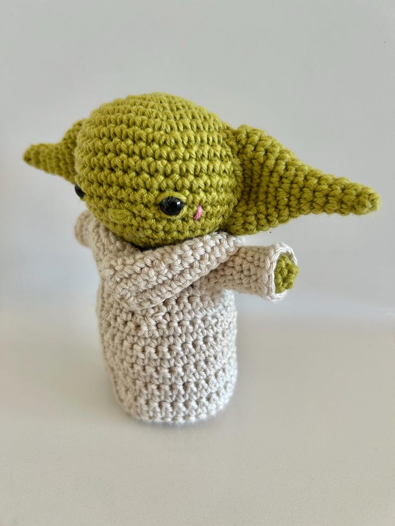Puede incluir: Mu&ntilde;eco de ganchillo hecho a mano de un personaje con orejas grandes y una t&uacute;nica beige. El mu&ntilde;eco tiene una cabeza verde, ojos negros y una peque&ntilde;a nariz rosa. La t&uacute;nica es de color beige claro y los brazos del mu&ntilde;eco est&aacute;n extendidos.