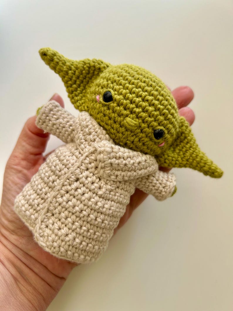 Puede incluir: Mu&ntilde;eca de ganchillo hecha a mano de un personaje con orejas grandes y una t&uacute;nica beige. La mu&ntilde;eca tiene ojos negros y mejillas rosadas. La mu&ntilde;eca est&aacute; hecha de hilo verde y beige.