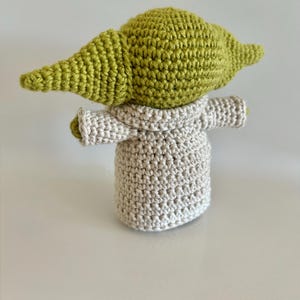 Puede incluir: Mu&ntilde;eca de ganchillo hecha a mano de un personaje con orejas grandes y cabeza verde claro. La mu&ntilde;eca lleva una t&uacute;nica gris claro. La mu&ntilde;eca est&aacute; sobre una superficie blanca.