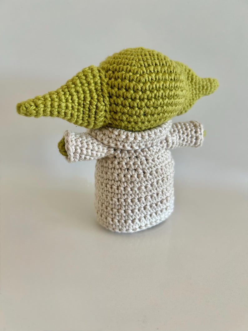 Puede incluir: Mu&ntilde;eca de ganchillo hecha a mano de un personaje con orejas grandes y cabeza verde claro. La mu&ntilde;eca lleva una t&uacute;nica gris claro. La mu&ntilde;eca est&aacute; sobre una superficie blanca.