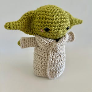 Puede incluir: Figura de ganchillo hecha a mano de un personaje con orejas grandes y ojos negros, con una t&uacute;nica beige. La figura es verde y beige, con un peque&ntilde;o rubor rosa en las mejillas. Aproximadamente 15 cm de altura.