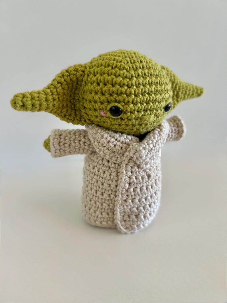 Puede incluir: Figura de ganchillo hecha a mano de un personaje con orejas grandes y ojos negros, con una t&uacute;nica beige. La figura es verde y beige, con un peque&ntilde;o rubor rosa en las mejillas. Aproximadamente 15 cm de altura.