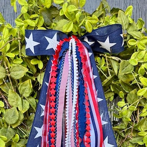 May include: A decorative wreath with a lush green base, adorned with a navy blue ribbon featuring white stars. Red, white, and blue ribbons cascade down, creating a patriotic display. The wreath is perfect for adding a touch of Americana to any door.