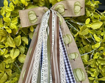 May include: A fall-themed wreath with a green leafy base. A burlap bow with green pumpkin illustrations is centered, with long ribbons in various patterns and colors, including brown, white lace, and checkered blue and white.