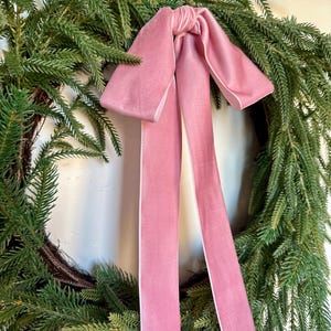 May include: A Christmas wreath made of evergreen branches, adorned with a large, light pink velvet bow. The bow's long, flowing ribbons add a touch of elegance to the festive decoration. The wreath is a classic holiday accent.