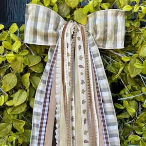 May include: A decorative wreath with a large bow made of tan and white striped fabric. Several ribbons in shades of brown, tan, and cream cascade down, some adorned with leaf patterns. The wreath is surrounded by green foliage.