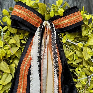 May include: A decorative black bow with orange and gold trim, accented with cascading ribbons in white, black, and gold. The bow is set against a backdrop of green foliage, creating a festive, seasonal display.