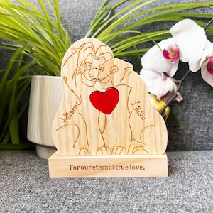 May include: Wooden lion figurine with a red heart cutout, featuring the names "Wilsery" and "Lillian". The base reads "For our eternal true love." The figurine is set against a backdrop of plants and flowers.