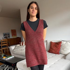 Può includere: Un abito senza maniche con scollo a V in tessuto a maglia marrone-rossastro. L'abito è indossato sopra una maglietta nera e abbinato a collant neri. Lo sfondo include un divano bianco, un tavolino nero e un tappeto fantasia.