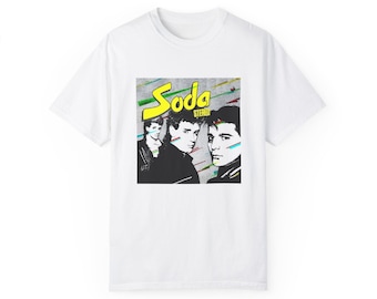 May include: White t-shirt featuring a graphic of three men with the word "Soda Stereo" in yellow and the word "Stereo" in white. The graphic has a retro design with colorful lines.