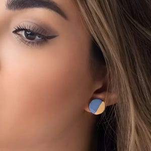 May include: A close-up of a person's eye and ear, with a blue and gold stud earring. The earring is a small, round disc, with a half-circle of blue and a half-circle of gold. The person has brown hair and is wearing eye makeup.