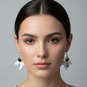 May include: White and blue floral drop earrings with gold-colored accents. The earrings feature a flower-shaped design with blue floral patterns and gold-colored metal details. The earrings are hanging from gold-colored hooks.