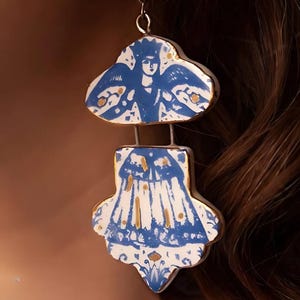 May include: Handmade ceramic earrings featuring a blue and white design with gold accents. The earrings have a cloud-like top and a fan-shaped bottom, connected by a silver wire. The design includes a figure with wings.