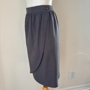 May include: A gray wrap skirt with a button closure. The skirt is displayed on a mannequin and has a draped design. The fabric appears to be a solid color and has a smooth texture. The skirt is knee-length.