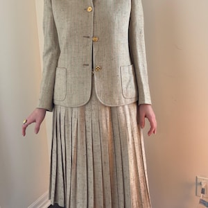 Puede incluir: Traje de falda de tweed beige con blazer y falda plisada. La blazer tiene botones dorados y bolsillos delanteros. La falda es hasta la rodilla. El traje tiene un sutil estampado moteado.