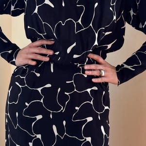 May include: A long-sleeved, black dress with a white floral pattern. The dress has a scoop neckline and a fitted waist. The skirt falls to the mid-calf. The model is wearing a silver ring. The dress is made of a lightweight fabric.