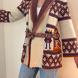 May include: Cream-coloured cardigan with geometric patterns in orange and navy blue. The cardigan features a shawl collar and a matching belt. The sleeves have a flared design, and the overall style is reminiscent of vintage knitwear.