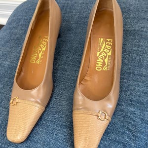 Peut inclure: Une paire d'escarpins Salvatore Ferragamo en cuir beige. Les chaussures ont un bout pointu avec un empiècement texturé et une boucle dorée. L'intérieur de la chaussure est beige avec le nom de la marque imprimé en or.