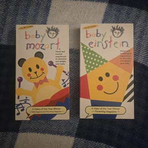 May include: Two VHS tapes, "Baby Mozart" and "Baby Einstein," are displayed. The "Baby Mozart" tape features a yellow bear playing a drum. The "Baby Einstein" tape has a smiling geometric face. Both tapes are labeled "A Video of the Year Winner."