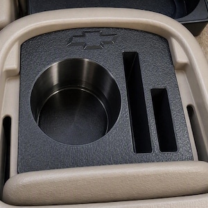 Puede incluir: Un inserto de consola central beige para automóvil con un portavasos circular de acero inoxidable y dos ranuras de almacenamiento rectangulares. El inserto de plástico negro texturizado muestra un logotipo Chevrolet en relieve.