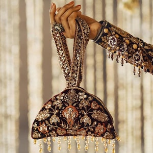 Op de afbeelding: Een bordeauxrode fluwelen handtas met ingewikkelde gouden borduursels en pailletten. De tas heeft een decoratief bloemmotief en een kralenfranje langs de onderkant. De handgreep is ook versierd met pailletten.
