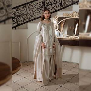 May include: A light beige formal outfit with intricate embroidery. The ensemble includes a long-sleeved tunic and wide-leg pants. The outfit features a sheer, flowing scarf and is accessorized with a gold necklace. The background includes a decorative mirror and staircase.