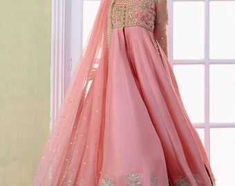 May include: A peach-coloured floor-length gown featuring a bodice with intricate gold embroidery and beadwork, long sheer sleeves, and a flared skirt. A matching sheer peach dupatta with gold sequin accents and a scalloped embroidered hem is draped over one shoulder.