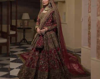 May include: A red and gold embroidered bridal gown with long sleeves and a full skirt. The gown features intricate detailing and a matching veil. The ensemble includes gold jewelry and a small purse. The dress is suitable for a wedding or special occasion.