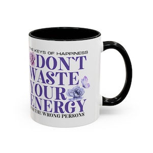 Könnte beinhalten: Ein weißer Keramikbecher mit schwarzem Henkel und schwarzem Innenraum. Die Außenseite zeigt violetten Text mit der Aufschrift THE KEYS OF HAPPINESS DON'T WASTE YOUR ENERGY FOR THE WRONG PERSONS, ergänzt durch Abbildungen violetter Blumen und eines Schmetterlings.